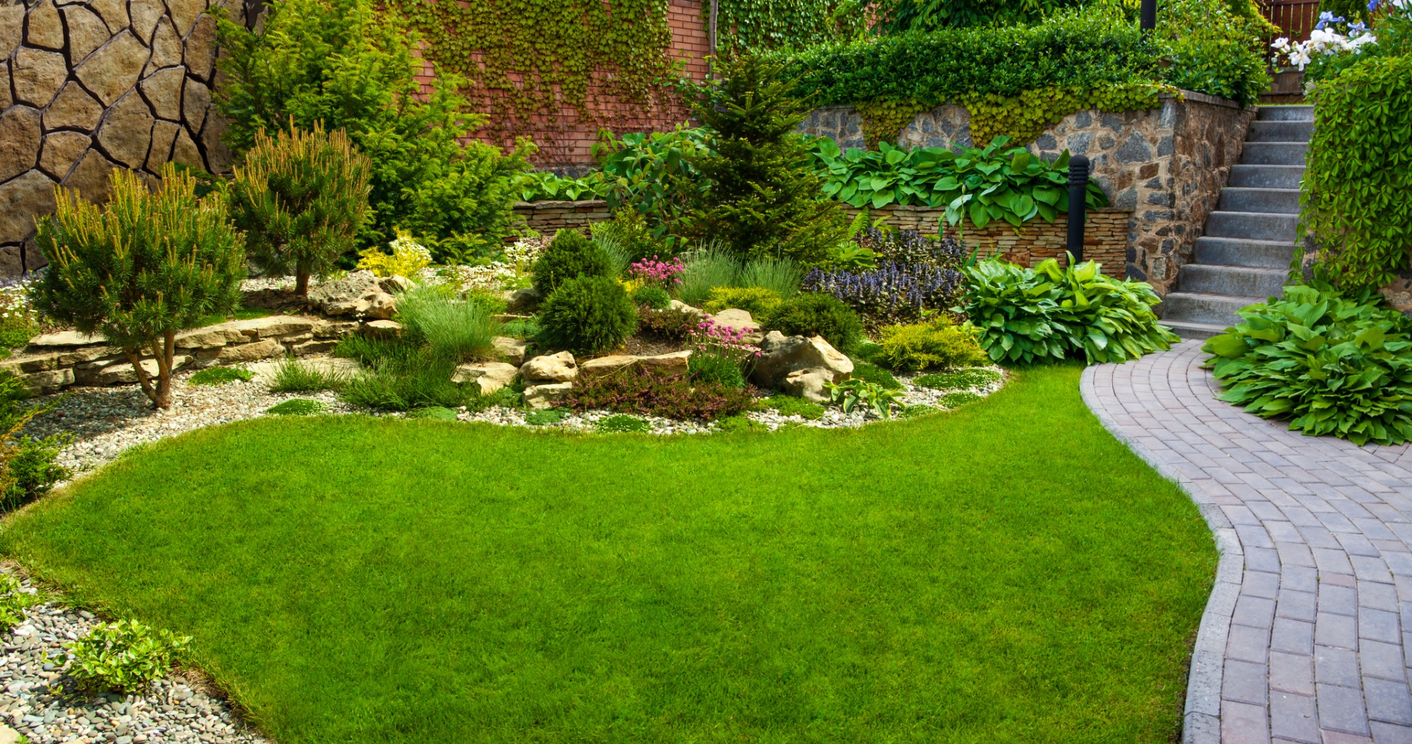 Well-manicured backyard garden with green lawn, curved paver pathway, ornamental plants, and ivy-covered stone walls