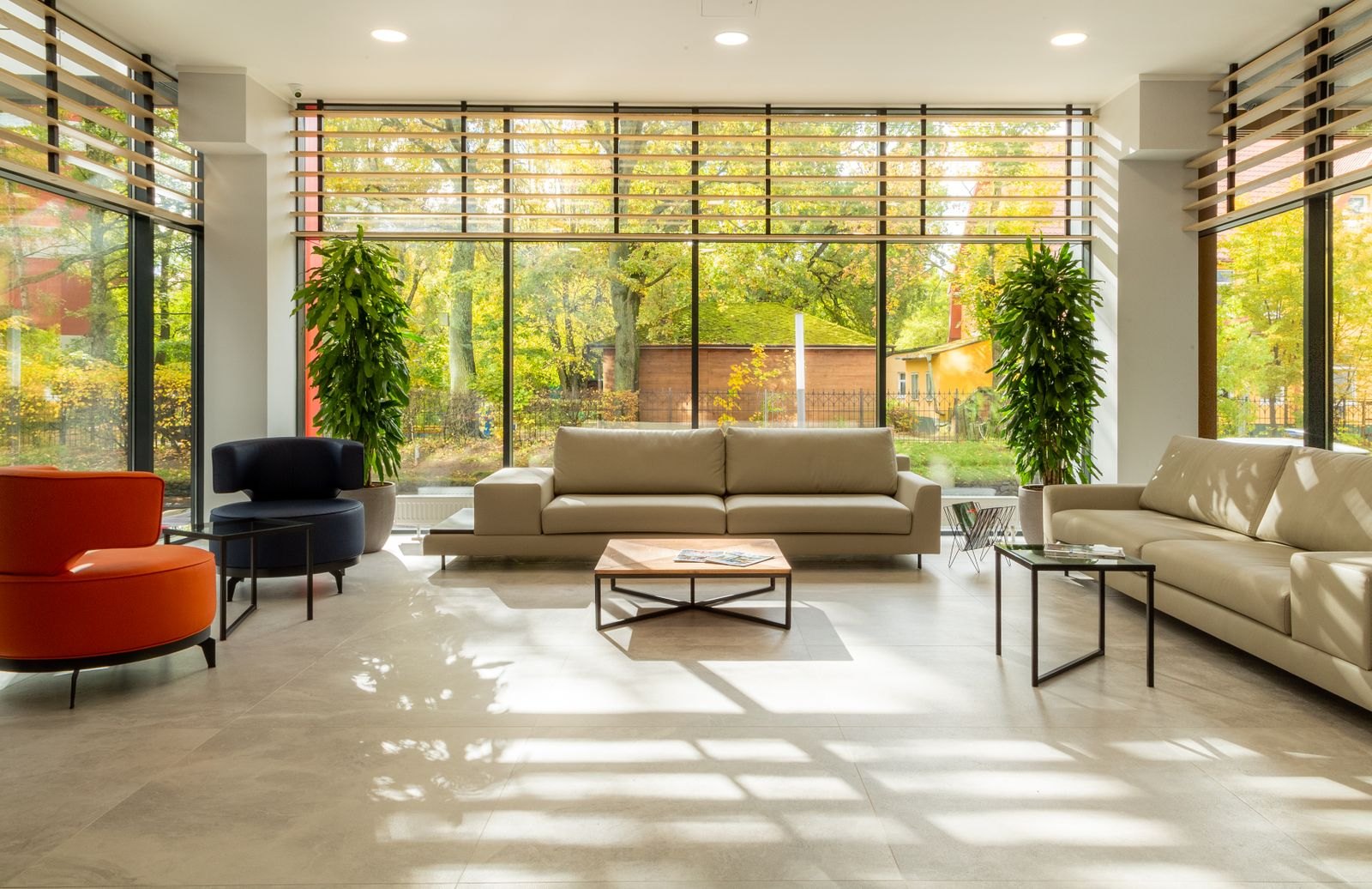 Modern lounge with beige sofas, red chairs, large windows overlooking autumn trees and brick buildings