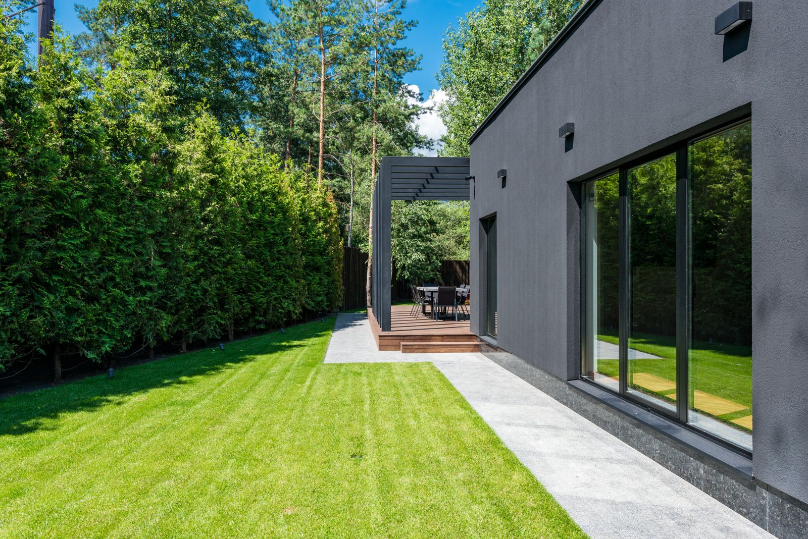 Modern gray house with large windows, concrete patio, wooden deck, and manicured lawn surrounded by green hedges and trees