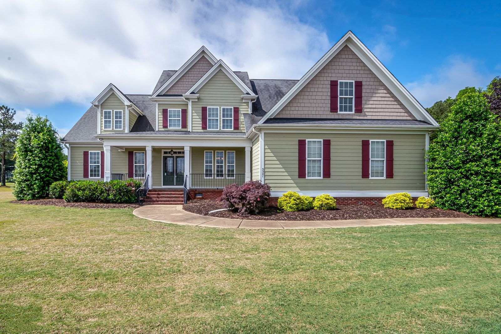 Two-story beige house with red shutters, brick foundation, front porch, and manicured landscaping