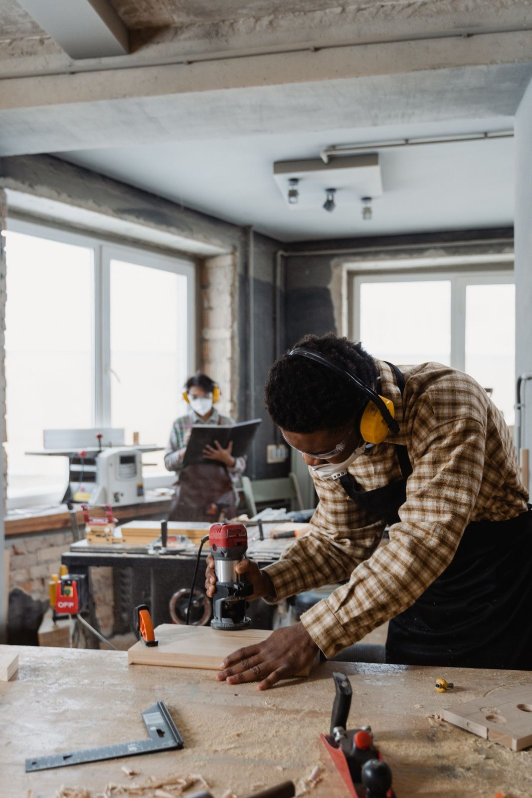 Carpenter using power tool on wood in workshop with colleague in background