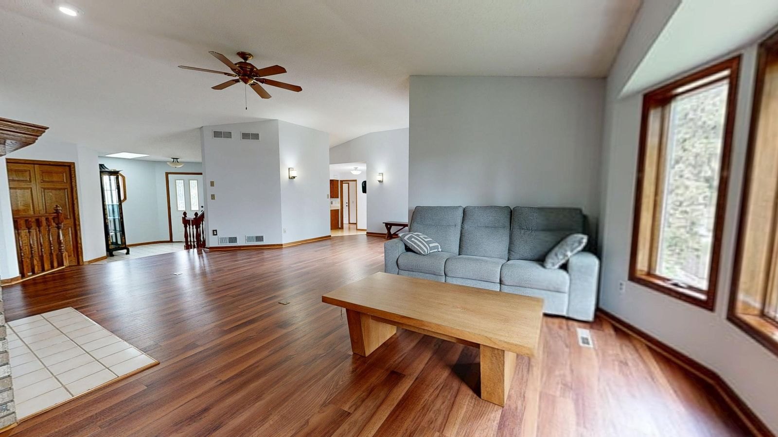 Spacious open-concept living room with hardwood floors, gray sofa, wooden table, and ceiling fan